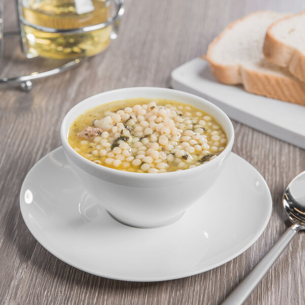 A bowl of soup with a Schonwald Grace white saucer on a table with bread and a spoon.