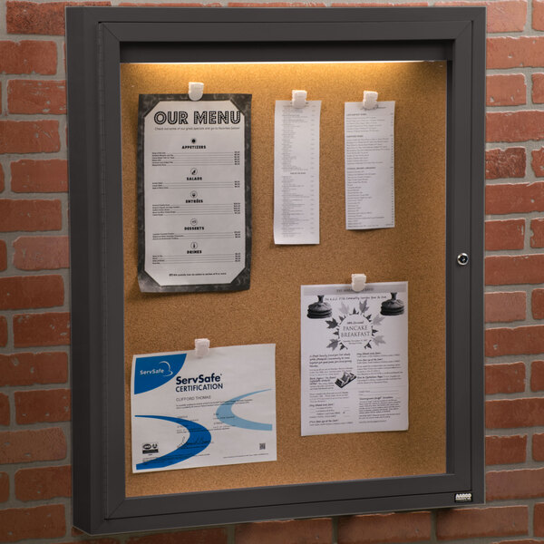 An Aarco bronze indoor lighted bulletin board cabinet with cork papers attached to it.