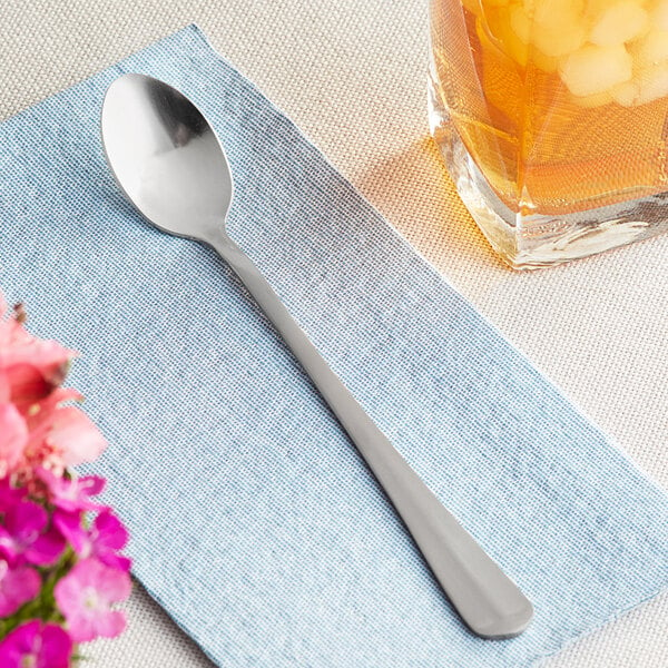 A Choice stainless steel iced tea spoon on a napkin next to a glass of liquid.