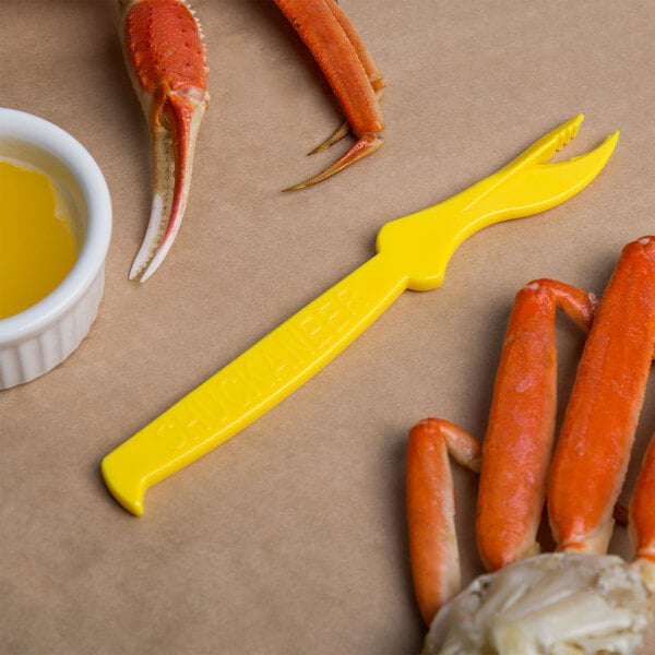 A crab claw with a yellow Choice Shuckaneer seafood sheller next to a bowl of crab legs.