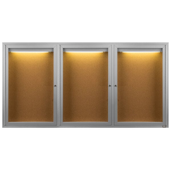 An Aarco satin anodized indoor bulletin board cabinet with three glass doors and lights.