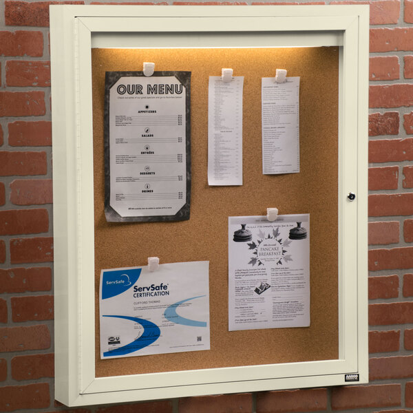 An ivory indoor bulletin board cabinet with a cork board inside with paper pinned to it.