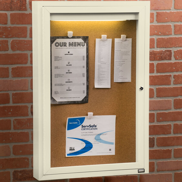 An ivory Aarco indoor lighted bulletin board cabinet with a white cork board and paper pinned to it.