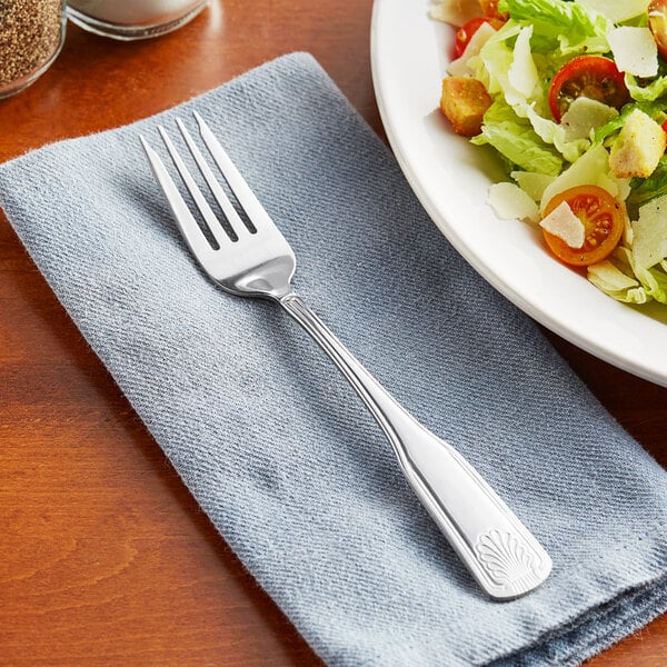 An Acopa stainless steel salad fork on a napkin next to a plate of salad.