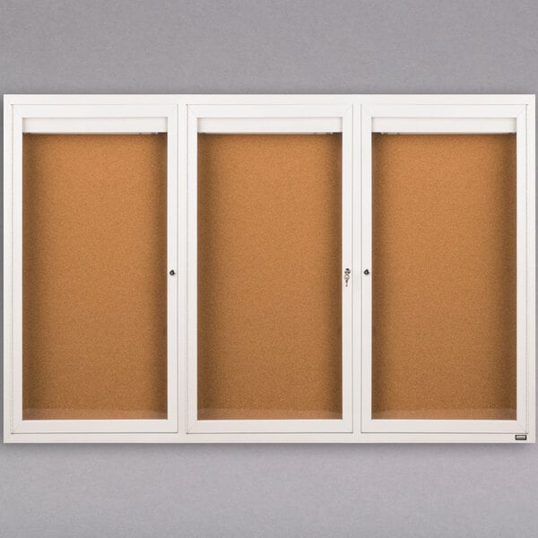 A white Aarco bulletin board cabinet with three glass doors.