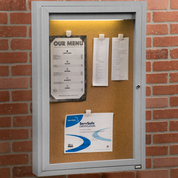 An Aarco satin anodized indoor lighted bulletin board cabinet with a white cork notice board on it.