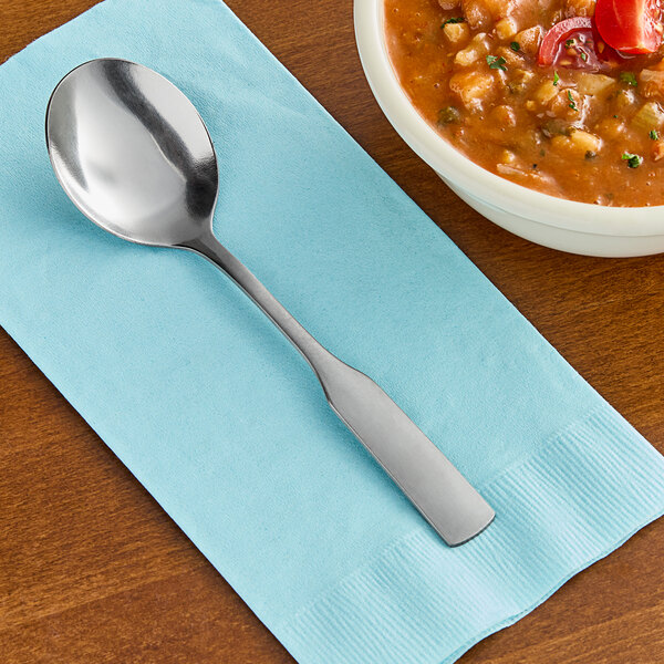 A Choice stainless steel bouillon spoon on a napkin next to a bowl of soup.