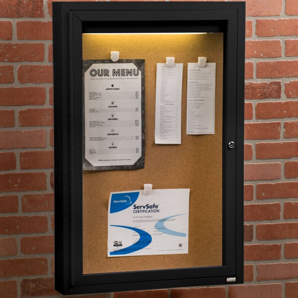 An Aarco black enclosed bulletin board cabinet with paper on it.