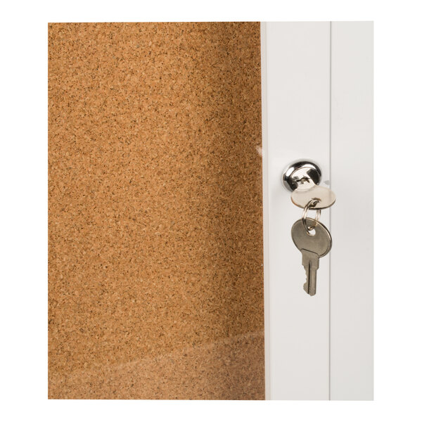 A close-up of a white-framed bulletin board cabinet with a cork surface and a lock with keys inserted.