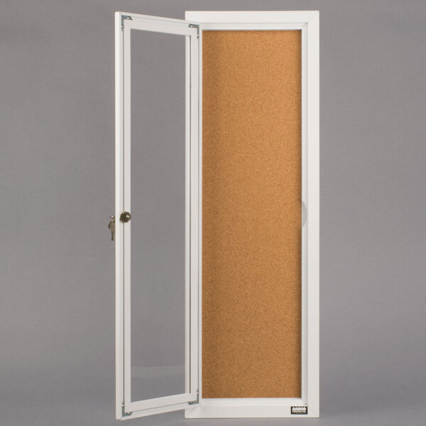 A white Aarco enclosed bulletin board cabinet with a cork board behind a clear glass door.