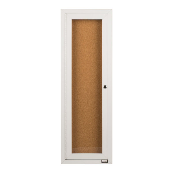A tall, narrow, white-framed enclosed bulletin board cabinet with a glass door and a corkboard interior.