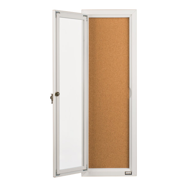 A tall, narrow bulletin board cabinet with a white frame, a corkboard interior, and a hinged glass door with a lock.