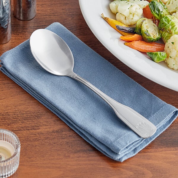 An Acopa stainless steel serving spoon on a napkin next to a plate of vegetables.