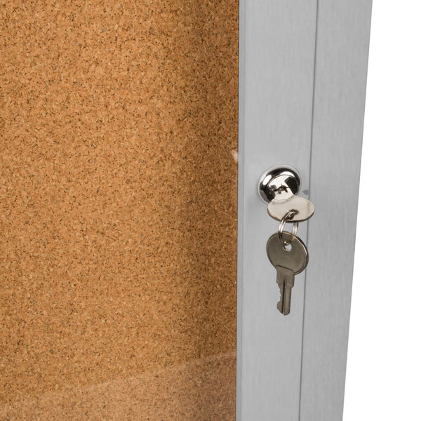 A satin anodized key in the door lock of an Aarco Bulletin Board Cabinet with cork board inside.