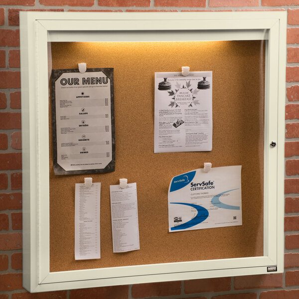 An ivory indoor enclosed bulletin board with a cork surface with papers attached.