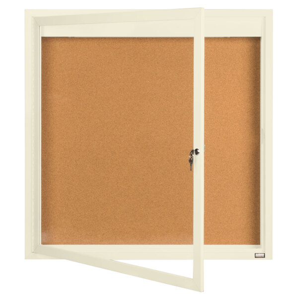 An ivory Aarco bulletin board cabinet with a cork board inside and a window with a key on it.