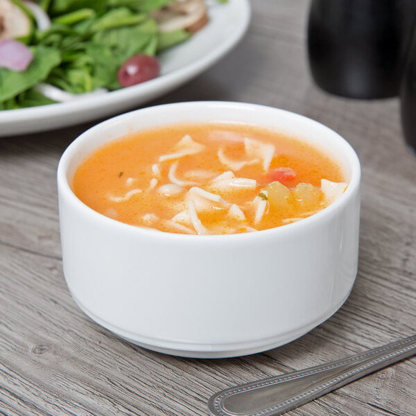 A Schonwald white porcelain bouillon cup filled with soup on a table with a plate of salad.