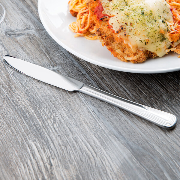 A Libbey stainless steel dinner knife with a fluted blade on a table with a plate of spaghetti, meat, and cheese.