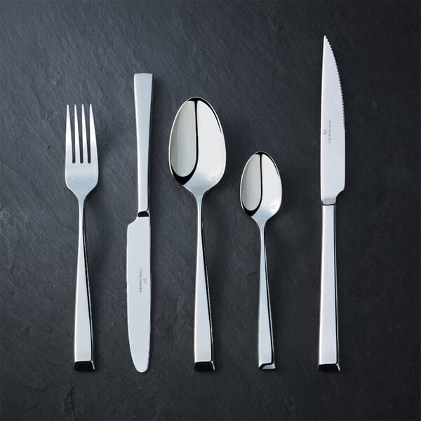Five pieces of shiny stainless steel cutlery, including a fork, two knives, and two spoons, are arranged side by side on a dark textured surface.