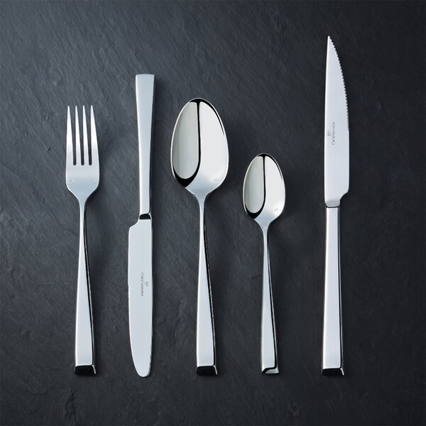 Five pieces of shiny stainless steel flatware, including a fork, two knives, and two spoons, are arranged side by side on a dark textured surface.