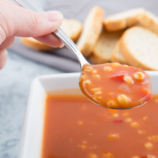 A hand holding a Libbey stainless steel bouillon spoon full of soup over a bowl of soup.