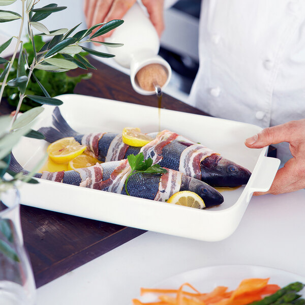 A person pouring oil onto fish wrapped in bacon in a white Villeroy & Boch rectangular baking dish.