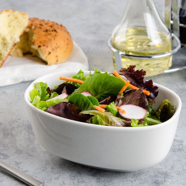 A Villeroy & Boch white porcelain oval bowl filled with salad on a counter with a bottle of oil and a bun.