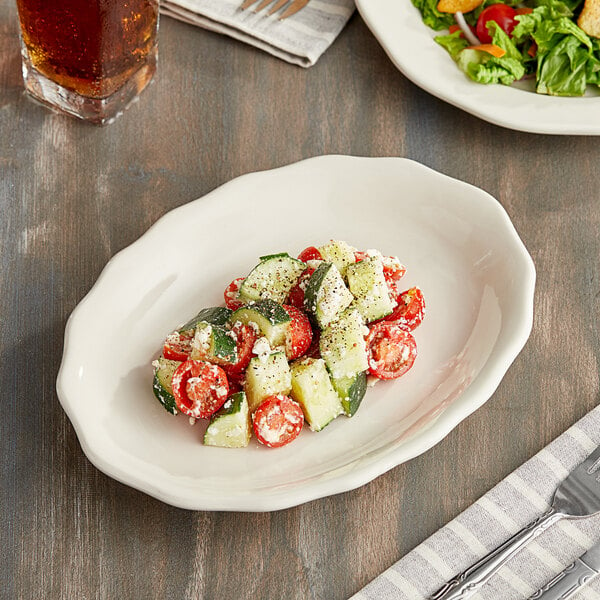 An Acopa ivory scalloped edge oval stoneware platter with a salad and drink on a table.