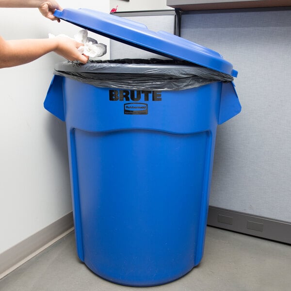 A woman putting a plastic bag into a blue Rubbermaid BRUTE trash can.