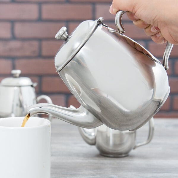 A hand pouring coffee into a silver Vollrath Orion teapot.