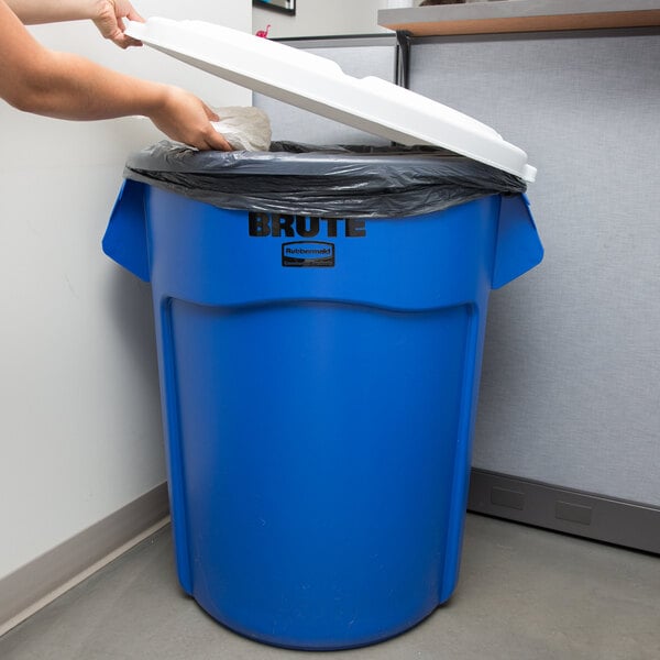 A woman putting a white lid on a blue Rubbermaid trash can.