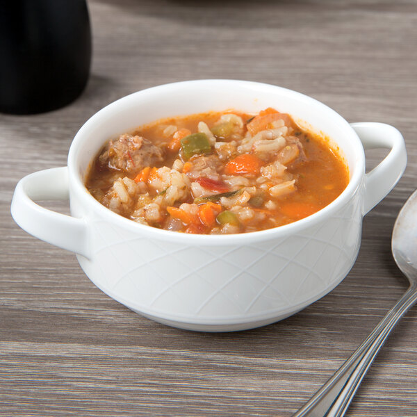 A white Villeroy & Boch porcelain soup cup with handles filled with soup on a table with a spoon.