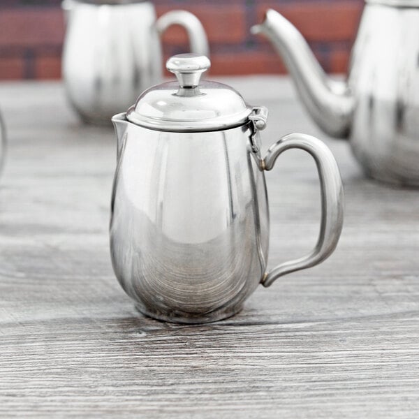 A Vollrath stainless steel creamer with a lid on a table.