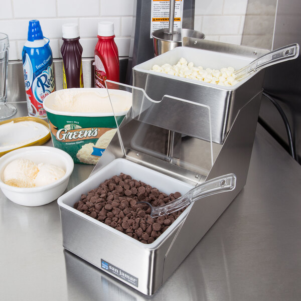 A San Jamar stainless steel condiment bar on a counter with bowls of food and a metal container of chocolate chips.