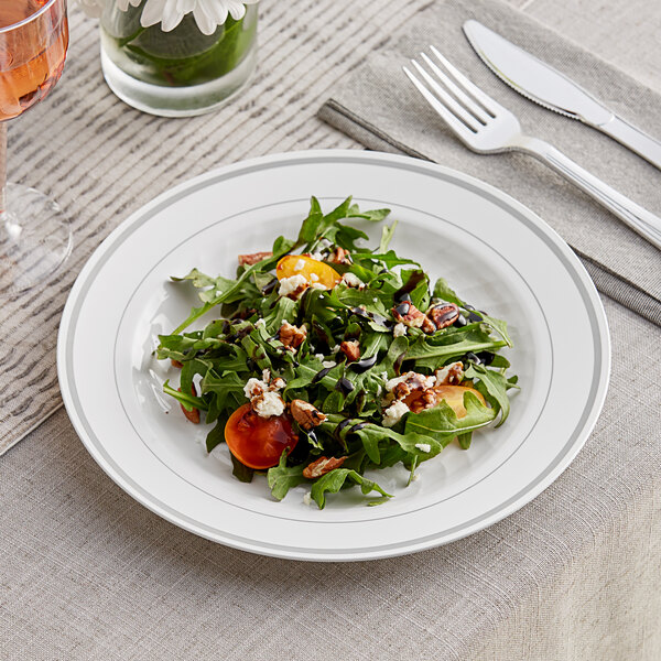 A WNA Comet white plastic plate with silver accent bands holding a salad with arugula, tomatoes, and feta cheese with a fork and knife on a table.