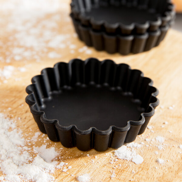 Two black Matfer Bourgeat fluted tartlet molds on a wooden surface.