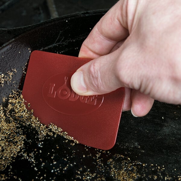 A person's hand holding a red rectangular Lodge pan scraper with a black background.