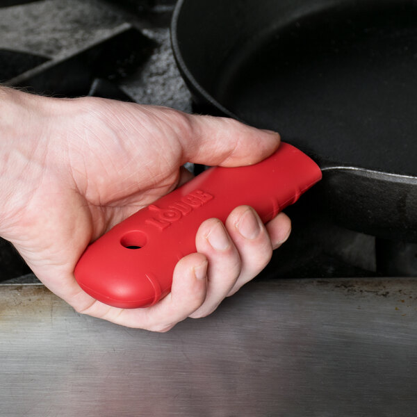 A person using a Lodge red silicone handle holder on a frying pan with a red handle.
