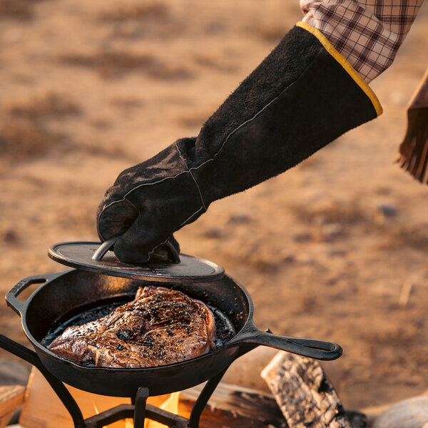 A person wearing Lodge leather gloves cooking steak on a cast iron skillet.