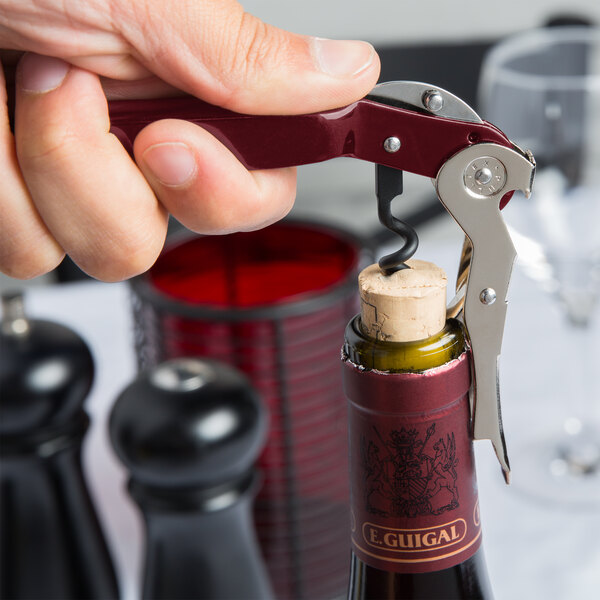 A hand holding a red Pullparrot Waiter's Corkscrew opening a wine bottle.