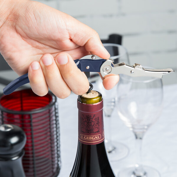 A hand uses a Pullparrot Waiter's Corkscrew with a blue handle to open a wine bottle on a table.