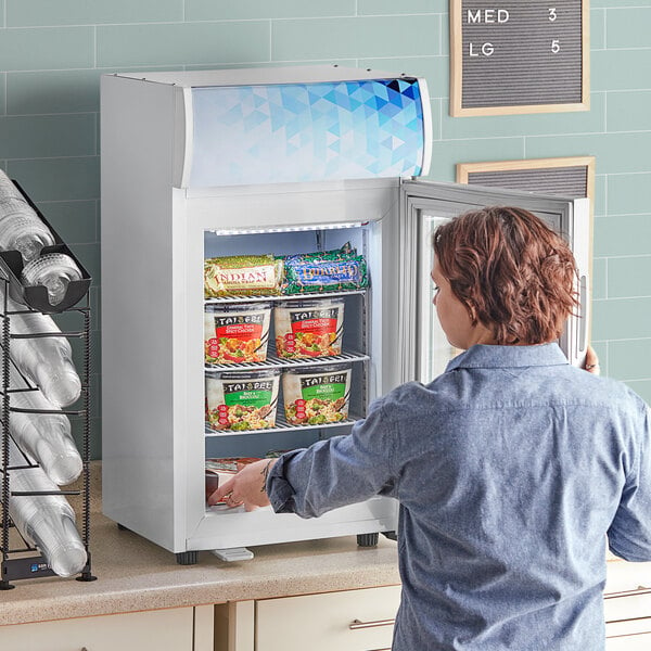 A woman opening the swing door on a white Avantco countertop freezer.