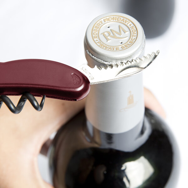 A person using a Franmara waiter's corkscrew with a burgundy handle to open a wine bottle on a table in a winery cellar.