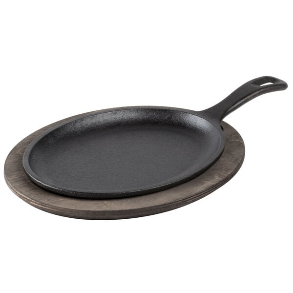 A Lodge pre-seasoned cast iron fajita skillet with handle on a wood underliner.