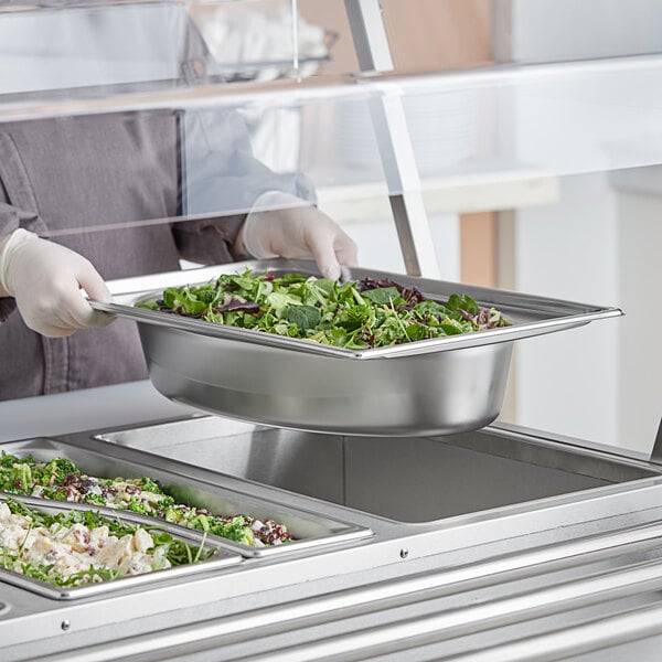 A hand holding a bowl of salad being served from a Vollrath stainless steel oval pan on a counter.