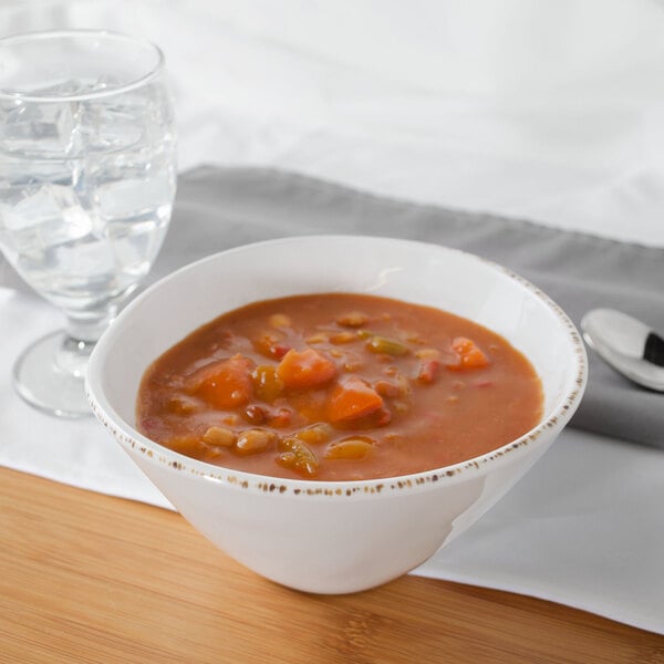 A white Urban Mill melamine bowl filled with soup and vegetables next to a glass of water.