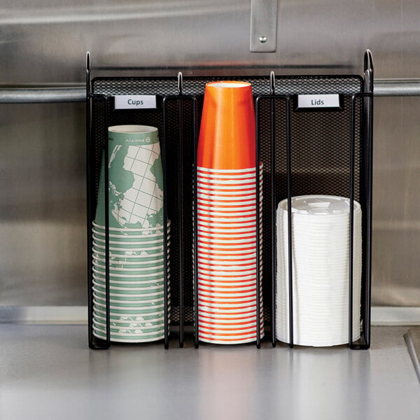 A Safco black steel mesh countertop cup and lid organizer holding cups on a counter.