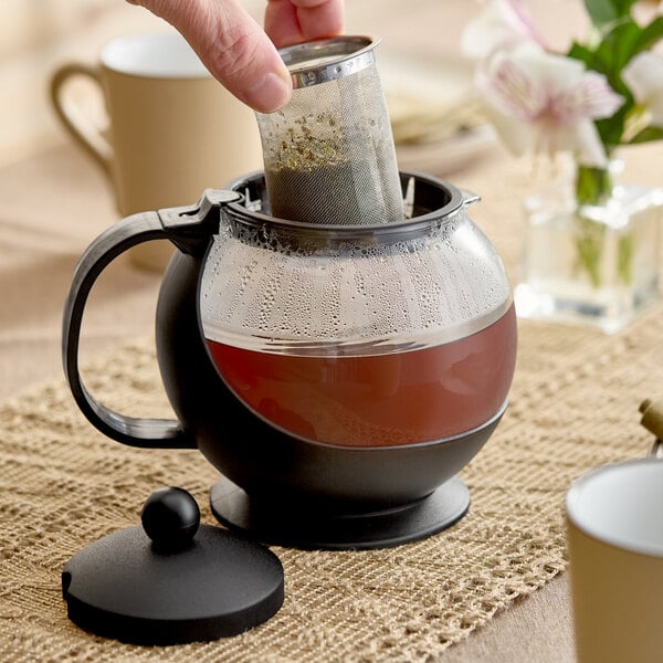 A glass teapot with a black handle and base, featuring a stainless steel infuser basket being lifted out.