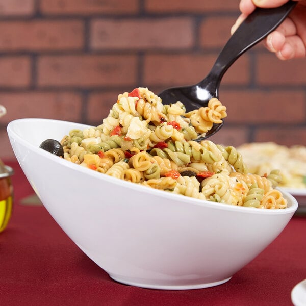 A hand holding a spoon of pasta over a white slanted catering bowl filled with pasta.