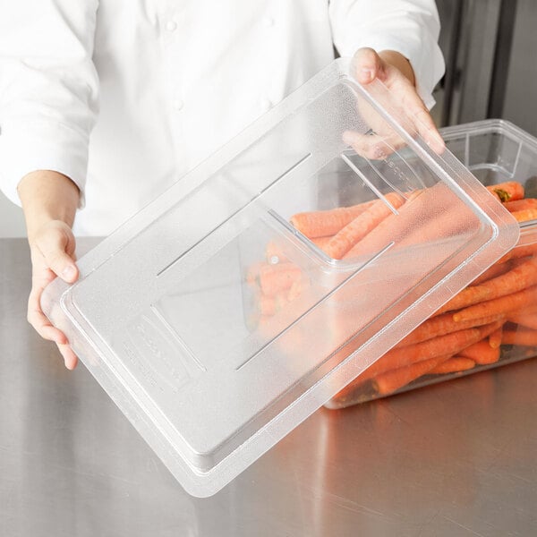A person holding a Rubbermaid clear polycarbonate food storage container full of carrots with a lid.